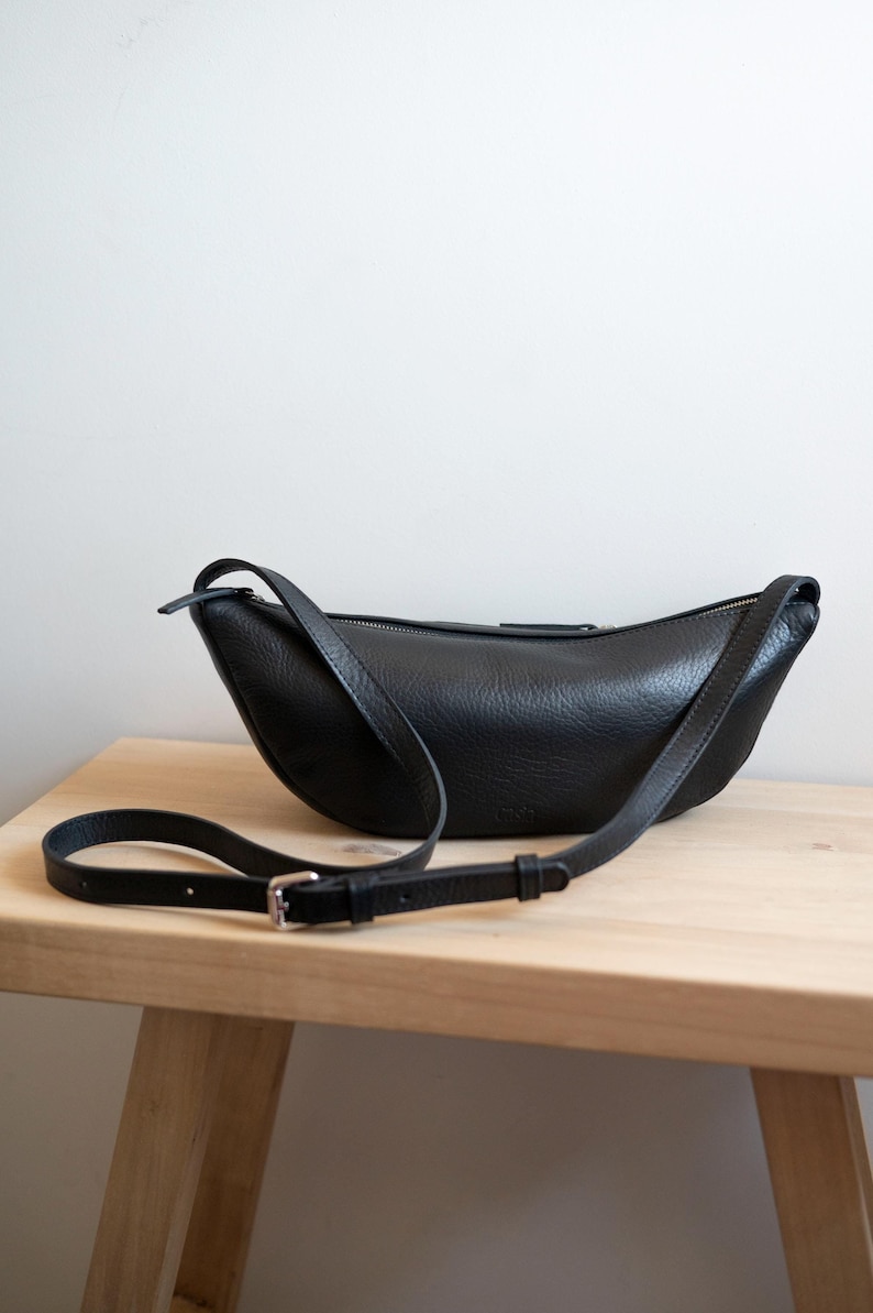 Black full grain leather banana bag on wooden table, moon shape vegetable tanned handmade crossbody sling bag product shot natural light