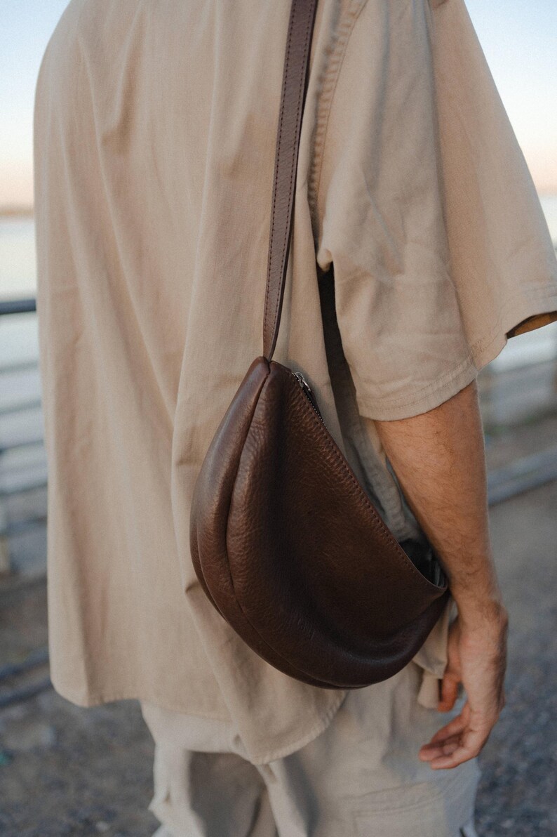 Man in shirt wearing chocolate full grain leather banana bag over shoulder, unisex vegetable tanned handmade moon shape crossbody sling bag