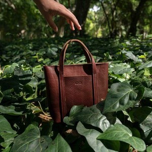 Hand holding bordeaux leather tote bag among green leaves in natural setting