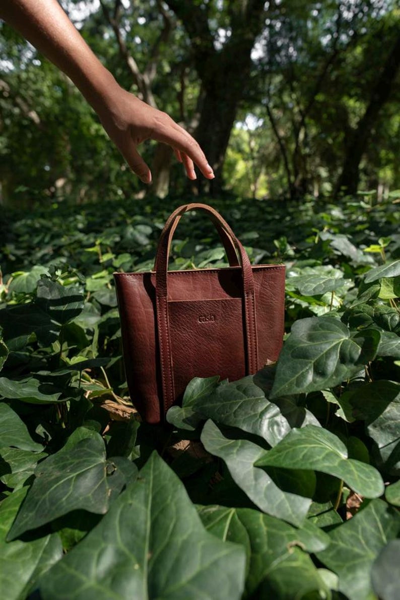Hand holding bordeaux leather tote bag among green leaves in natural setting