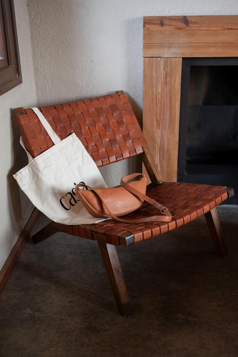 Tan full grain leather banana bag with Casia canvas tote on woven leather chair by fireplace, handmade vegetable tanned sling bag lifestyle interior