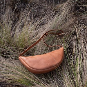 Tan handmade leather banana bag lying on dry pampas grass, moon shape full grain vegetable tanned crossbody sling bag, natural leather texture visible