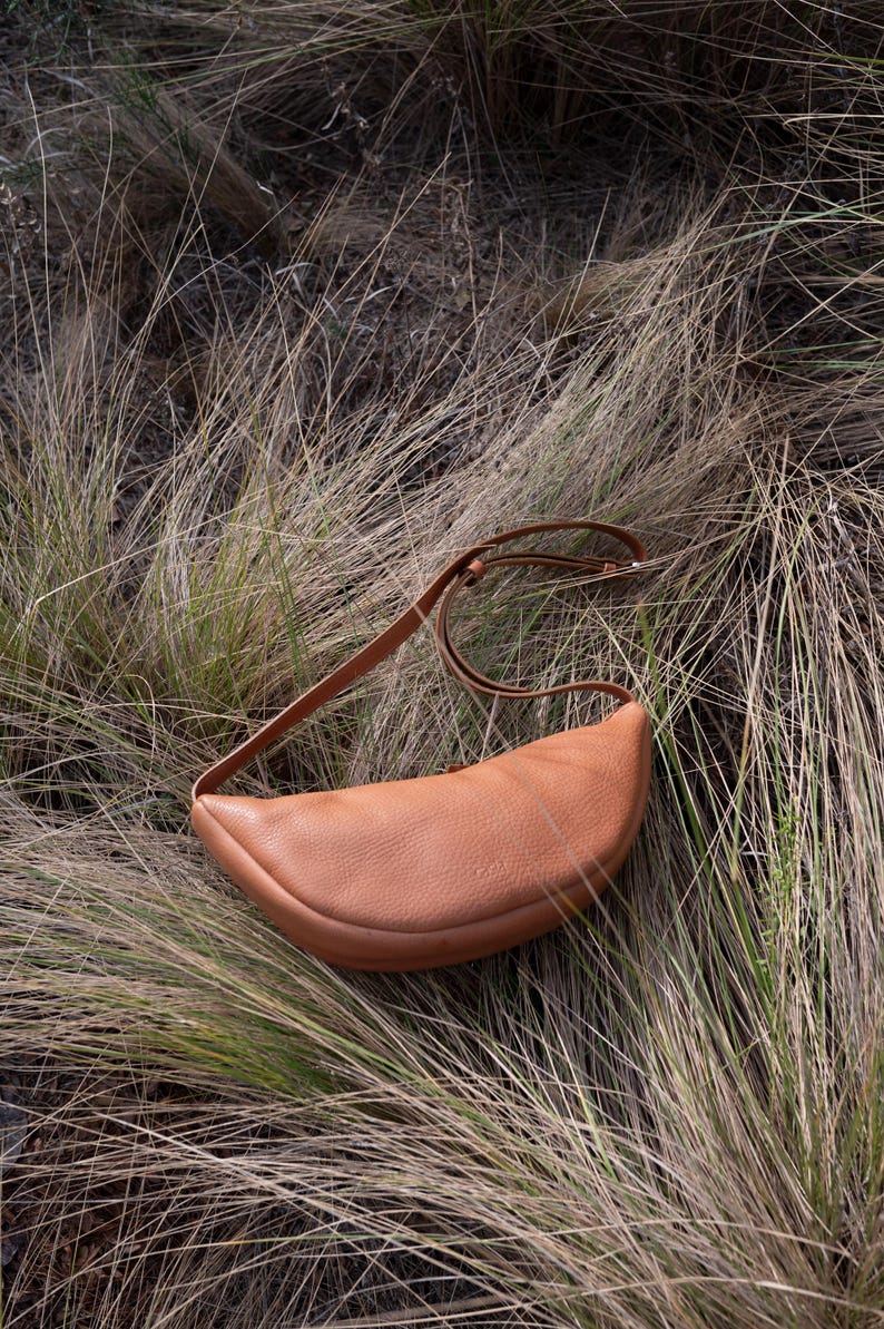 Tan handmade leather banana bag lying on dry pampas grass, moon shape full grain vegetable tanned crossbody sling bag, natural leather texture visible