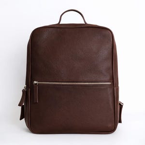 Puede incluir: Una mochila de cuero marrón con forma rectangular. Cuenta con un bolsillo frontal con cremallera, un asa superior y correas ajustables. La bolsa tiene una cremallera plateada y un diseño minimalista.