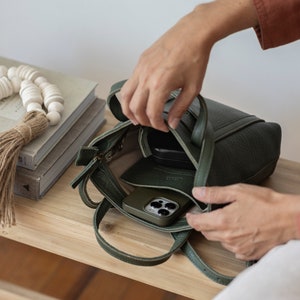 Hands opening olive green leather bag on wooden table showing zipper closure