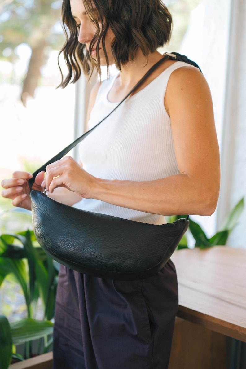 Woman opening black full grain leather banana bag showing interior, handmade vegetable tanned crossbody sling bag with zip closure lifestyle