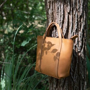 Woman holding tan leather tote bag near tree in natural outdoor setting
