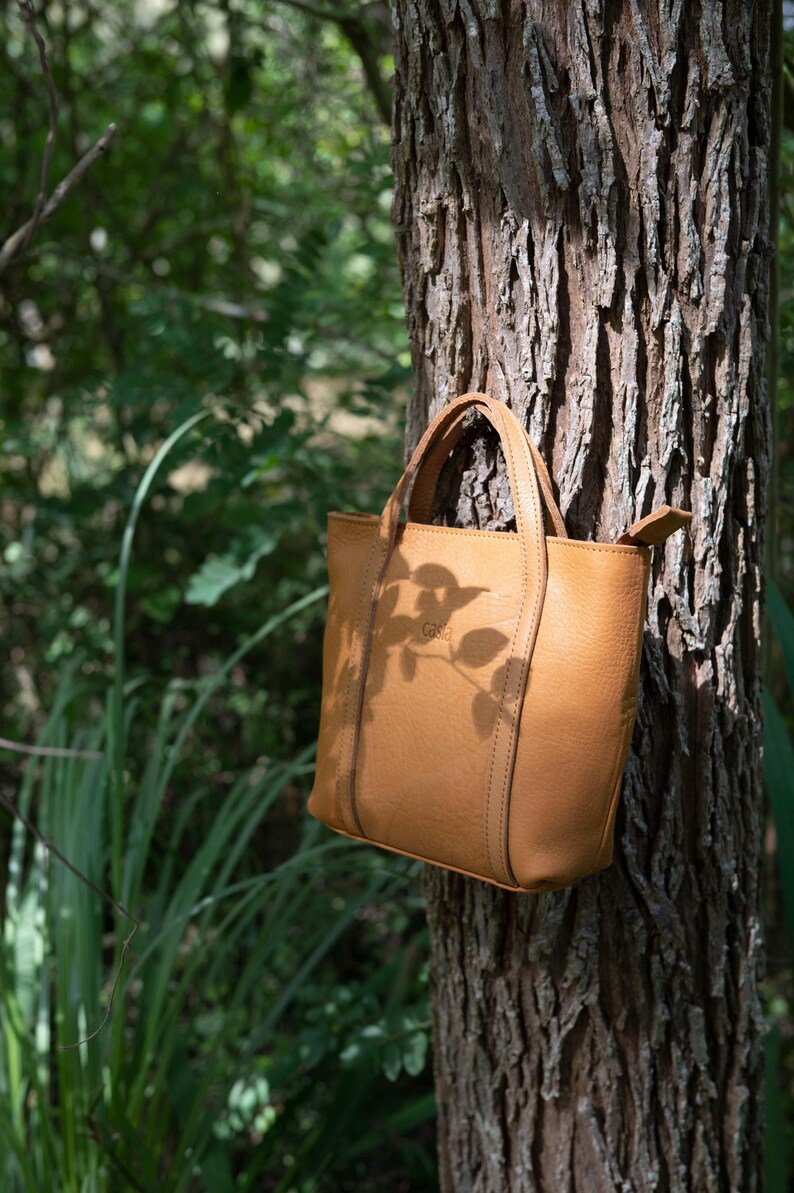 Woman holding tan leather tote bag near tree in natural outdoor setting
