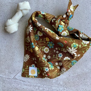 May include: A brown dog bandana with a floral pattern featuring white, blue, and yellow flowers. A white dog bone is next to the bandana. The bandana has a small tag that says "pretty snappy".