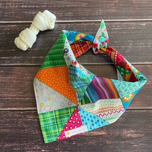 May include: A colorful patchwork dog bandana with a blue button and the text "mostly sunny" in a yellow circle. The bandana is tied in a knot and is on a wooden background.