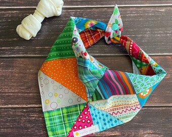 May include: A colorful patchwork dog bandana with a blue button and the text "mostly sunny" in a yellow circle. The bandana is tied in a knot and is on a wooden background.