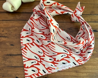 May include: A white and red ghost-patterned dog bandana with a button closure. The bandana is folded and lies on a wooden surface next to two white dog treats. The bandana has the text "mostly sunny".