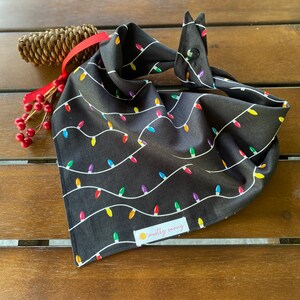 May include: Black bandana with a white string of colorful lightbulbs pattern. The bandana has a snap closure and a small yellow tag with the text "mostly sunny".