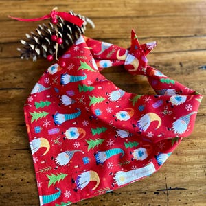 May include: Red Christmas-themed dog bandana with a pattern of gnomes, Christmas trees, and snowflakes. The bandana is tied in a knot and is next to a pinecone with red berries.