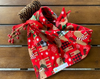 Op de afbeelding: Rode bandana met een kerstmotief van elanden en een wit sneeuwvlokkenpatroon. De bandana heeft een drukknopsluiting en een "mootly mommy"-label.