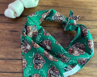 May include: Green dog bandana with a football-themed pattern featuring football players and play diagrams. The bandana has a tie closure and is next to a white dog bone. The bandana has the text "mostly sunny".