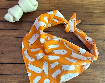 May include: Orange dog bandana with a white ghost pattern. The bandana is tied in a knot. Two white dog treats are in the upper left corner. The bandana has the text "mostly sunny" on it.