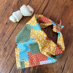 May include: A patchwork dog bandana with a floral and geometric pattern in yellow, blue, green, and orange. The bandana is tied with a button closure. Two white dog treats are next to the bandana. The text "mostly sunny!" is on the bandana.
