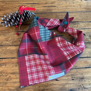 May include: A red plaid dog bandana with a patchwork design. The bandana features various red, white, and green plaid patterns. It has a button closure and a small tag with the text "mostly sunny".