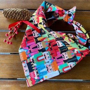 May include: A colorful bandana with a pattern of nutcracker soldiers. The bandana is tied in a knot and has a small tag that says "mostly sunny".