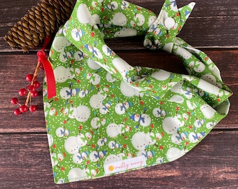 Op de afbeelding: Groene bandana met een sneeuwmanmotief en witte sneeuwvlokken. De bandana heeft een rode en witte knoopsluiting en een "mostly sunny"-label.