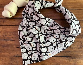 May include: A black and white Halloween dog bandana with a repeating ghost pattern. The bandana is tied in a knot. Two white dog treats are next to the bandana. The bandana has the text "mostly sunny".