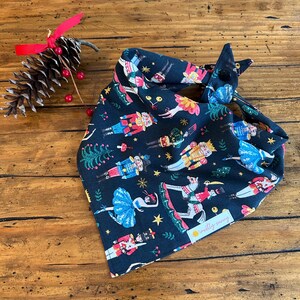 May include: A black bandana with a festive Nutcracker and ballerina print. The bandana features a repeating pattern of Nutcrackers, ballerinas, and Christmas trees. The bandana has a small yellow tag with the text "mostly sunny" on it.
