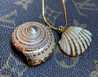 Puede incluir: Dos collares de oro con colgantes de conchas marinas. Un colgante es una concha blanca con detalles dorados, el otro es una concha marrón y blanca con detalles dorados.
