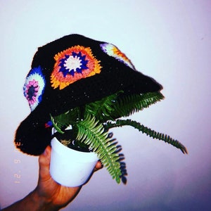 Può includere: Un cappello a secchiello in crochet nero con motivi colorati a quadrati della nonna. Il cappello è tenuto da una mano e una pianta di felce verde cresce dalla cima del cappello.