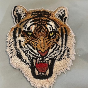 Könnte beinhalten: Gestickter Tigerkopf-Aufnäher mit detaillierten Nähten. Der Tiger hat einen wilden Ausdruck mit offenem Mund, der das rote Innere des Mundes und scharfe Zähne zeigt. Der Aufnäher hat einen cremefarbenen Rand und ist vor einem hellblauen Hintergrund platziert.