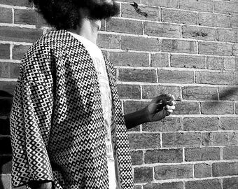May include: Black and white image of a person wearing a patterned kimono-style jacket over a white t-shirt. The jacket features a geometric design. The person is standing in front of a brick wall, wearing sunglasses and has a full beard.