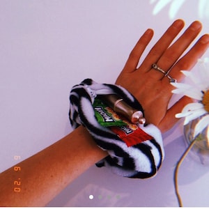 May include: A black and white zebra-print wristlet with a zipper closure. The wristlet is holding a small silver tube and two snack-sized bags of chips. The bags are labeled "MAC'n" and "MOM'N".