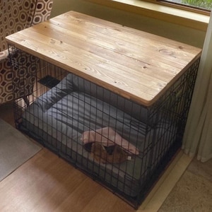 May include: A rectangular black wire pet crate featuring a smooth, light brown wooden top surface. Inside the crate, a gray rectangular pet bed is placed on the floor, with a small, folded pink fabric item resting on top of the bed surface.