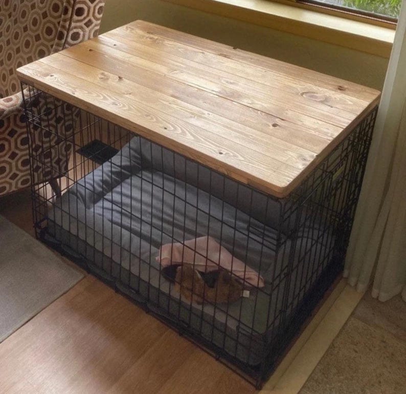 May include: A rectangular black wire pet crate featuring a smooth, light brown wooden top surface. Inside the crate, a gray rectangular pet bed is placed on the floor, with a small, folded pink fabric item resting on top of the bed surface.