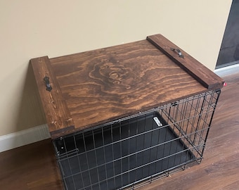 Può includere: Una gabbia per cani con un piano in legno marrone scuro e una gabbia in metallo nero. Il piano in legno ha due maniglie in metallo scuro. La gabbia è su un pavimento marrone scuro.