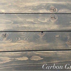 May include: Close-up of a wood surface stained in a dark gray color called "Carbon Gray". The wood has a natural grain pattern and knots.