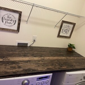 May include: A white laundry room with a dark wood countertop over a white washing machine and dryer. The walls are white and there are two framed signs on the wall. The sign on the left says "The Laundry Room Do It Yourself" and the sign on the right says "The laundry is looking at me Dirty Again".