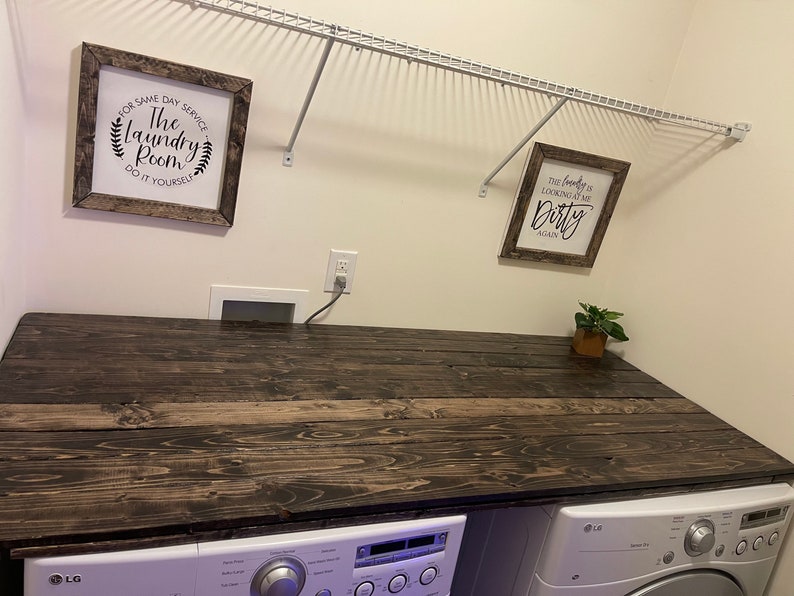May include: A white laundry room with a dark wood countertop over a white washing machine and dryer. The walls are white and there are two framed signs on the wall. The sign on the left says "The Laundry Room Do It Yourself" and the sign on the right says "The laundry is looking at me Dirty Again".