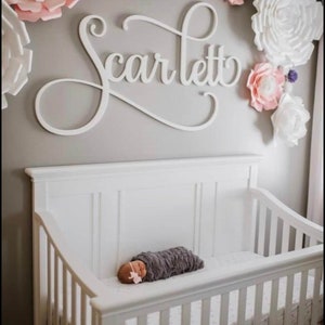 May include: A white wooden sign with the name "Scarlett" in cursive script hangs above a white crib with a baby sleeping inside. The wall is decorated with large paper flowers in shades of pink, white, and purple.