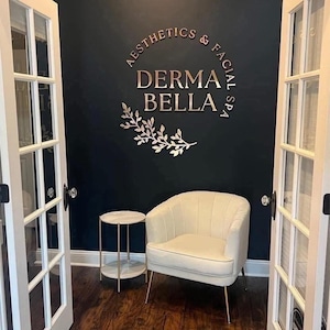 May include: A wall-mounted sign with metallic gold lettering reading DERMA BELLA AESTHETICS & FACIAL SPA, accompanied by a gold leaf branch graphic. Below the sign sits a cream-colored upholstered armchair and a small round side table.
