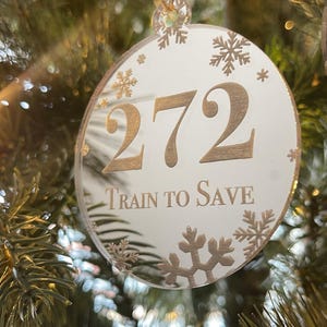 Könnte beinhalten: Ein runder, weißer Weihnachtsschmuck mit goldenem Text und Schneeflocken-Akzenten. Der Schmuck trägt die Aufschrift "272 Train to Save". Er hängt an einer transparenten, dekorativen Schnur vor einem verschwommenen Weihnachtsbaumhintergrund.