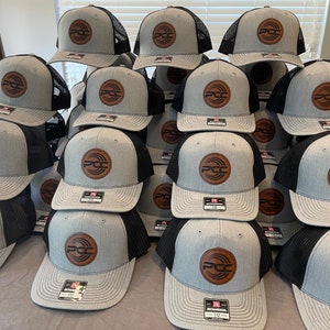 May include: A stack of gray and black trucker hats with a brown leather patch featuring the letters "PCC" inside a circular design. The hats have a curved brim and a mesh back. The hats are stacked on top of each other.