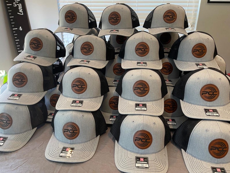 May include: A stack of gray and black trucker hats with a brown leather patch featuring the letters "PCC" inside a circular design. The hats have a curved brim and a mesh back. The hats are stacked on top of each other.