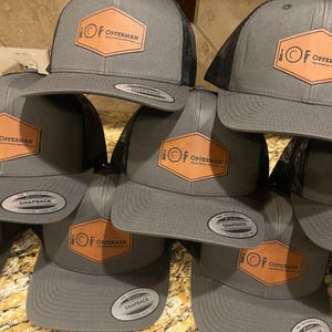 May include: A group of six gray and black trucker hats with a brown leather patch on the front. The patch features a stylized fork and spoon with a circle in the middle and the text "OF OPFERMAN" in black lettering. The hats have a black mesh back and a white "SNAPBACK" label.
