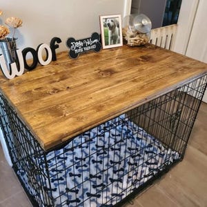 May include: A black wire dog crate with a rectangular stained wood top. The crate features a white blanket with a black bone pattern inside. On the wood surface sits a metal vase with paper flowers, a black sign reading Woof, a bone-shaped sign reading Dogs Welcome People Tolerated, a framed photo, and a glass jar.