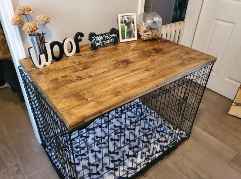 May include: A black wire dog crate with a rectangular stained wood top. The crate features a white blanket with a black bone pattern inside. On the wood surface sits a metal vase with paper flowers, a black sign reading Woof, a bone-shaped sign reading Dogs Welcome People Tolerated, a framed photo, and a glass jar.