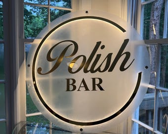May include: A round, illuminated sign with the words "Polish BAR" in gold lettering. The sign is mounted on a glass table in front of a window. The sign has a frosted appearance with a black border and a warm, inviting glow.