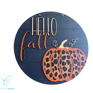 以下が含まれることがあります： 「Hello Fall」という金色の文字と、大きくオレンジ色、茶色、黒色のヒョウ柄の南瓜が描かれた丸い木製の看板です。