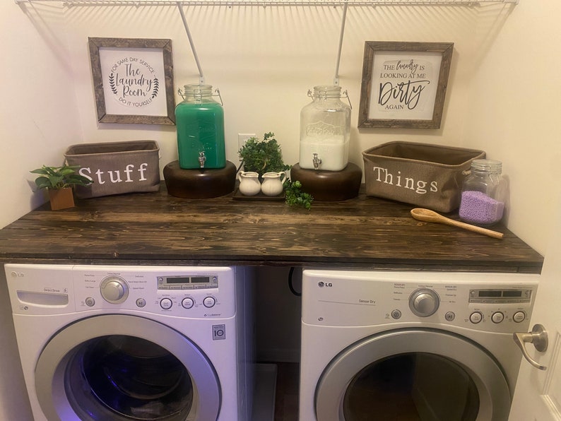 May include: A laundry room with two white washing machines, a wooden countertop, two glass dispensers with green and white liquids, two baskets labeled "Stuff" and "Things", and two signs that say "The Laundry Room Do It Yourself" and "The Laundry is Looking at Me Dirty Again".