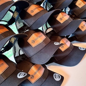 May include: A collection of black trucker hats with a brown leather patch featuring the text "CONSTRUCTION COMPONENTS" and a logo. The hats have a mesh back and a curved brim. The hats are stacked on top of each other.